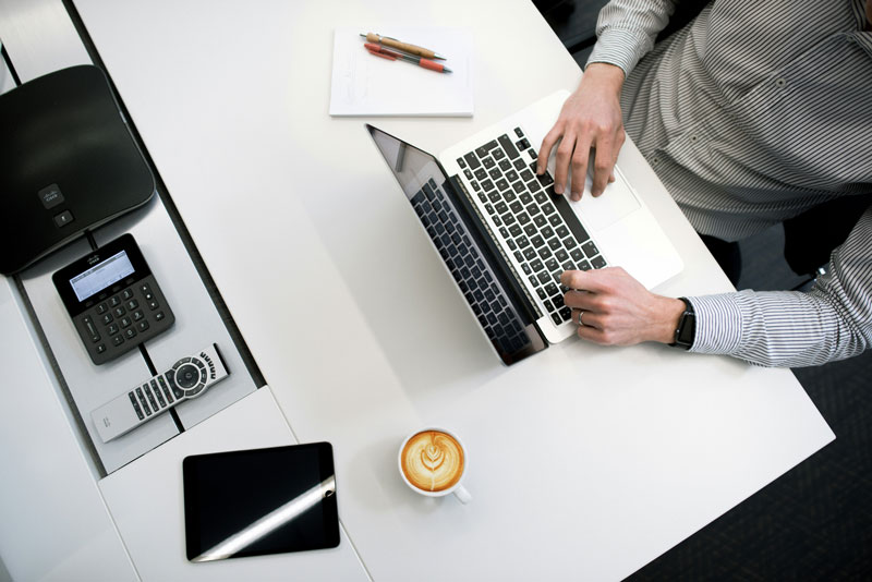 Vista de cima para baixo de uma mesa de escritório com um homem usando um laptop, próximo a um tablet com tela preta, um telefone, uma xícara de café com arte de latte e anotações, simbolizando o trabalho de tributação para empresas estrangeiras.